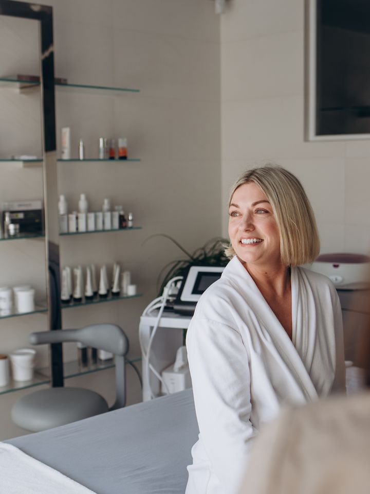 A person in a robe seated in a treatment room lined with skincare shelves.