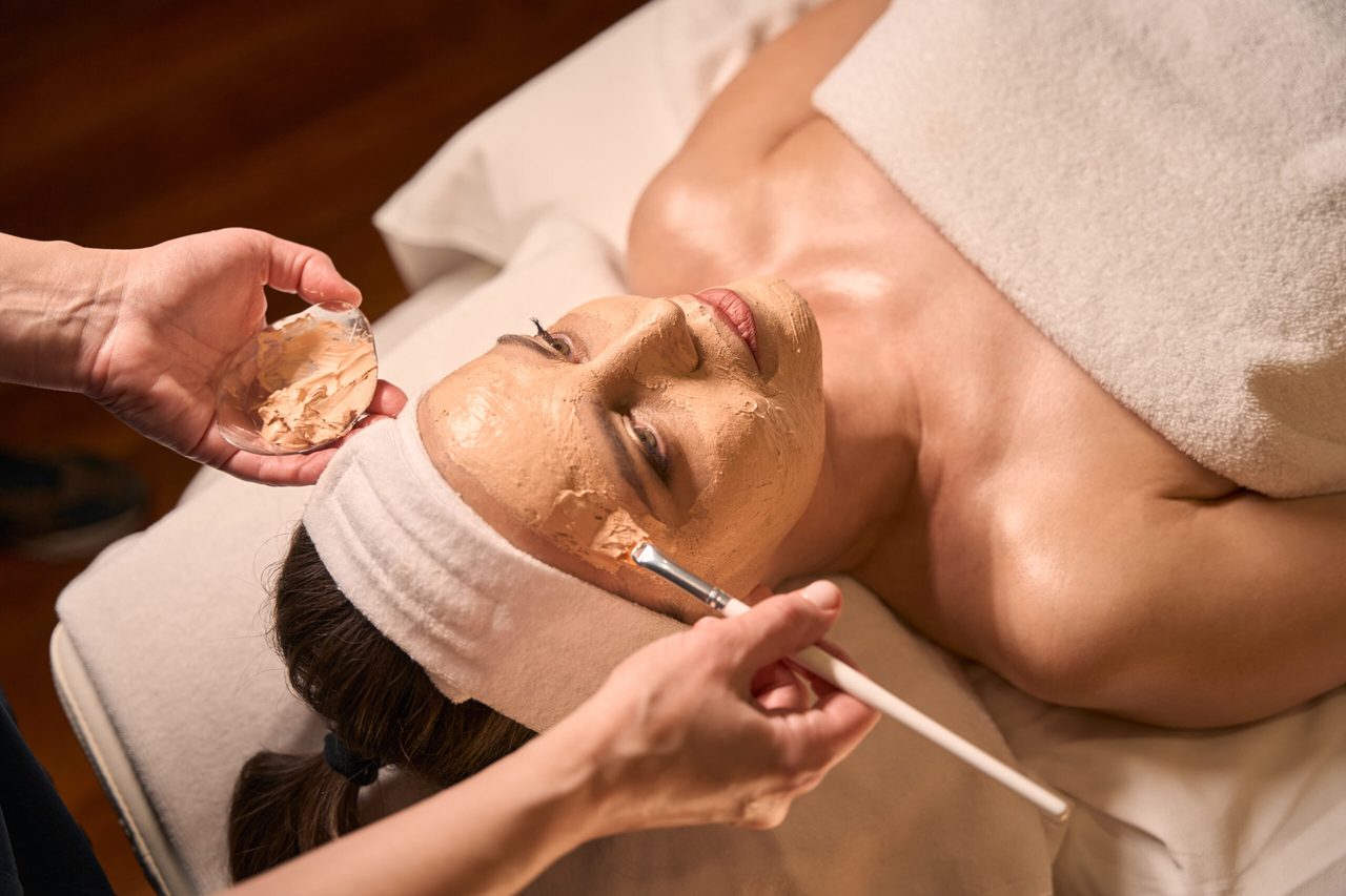 A clay treatment mask being applied to the cheek with a brush.